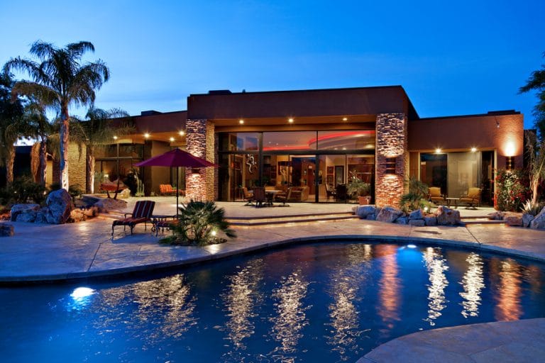 A luxurious modern home with large glass windows is beautifully illuminated at dusk, showcasing a spacious patio with seating areas, palm trees, and a curved swimming pool reflecting the warm exterior lighting.
