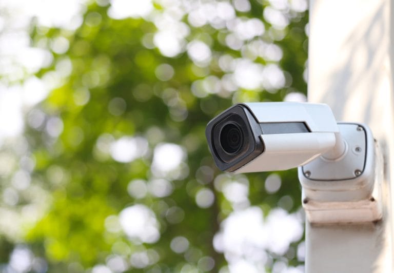 Close-up of a modern outdoor security camera mounted on a white wall, with a blurred green leafy background in daylight.