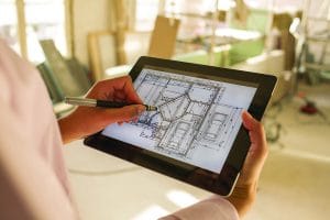 A person uses a stylus on a tablet displaying a detailed architectural floor plan while standing in a construction or renovation site.