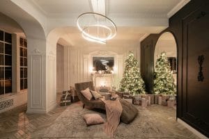 An elegant living room decorated for Christmas with two lit trees, a cozy chaise lounge, wrapped gifts, and modern circular lighting.