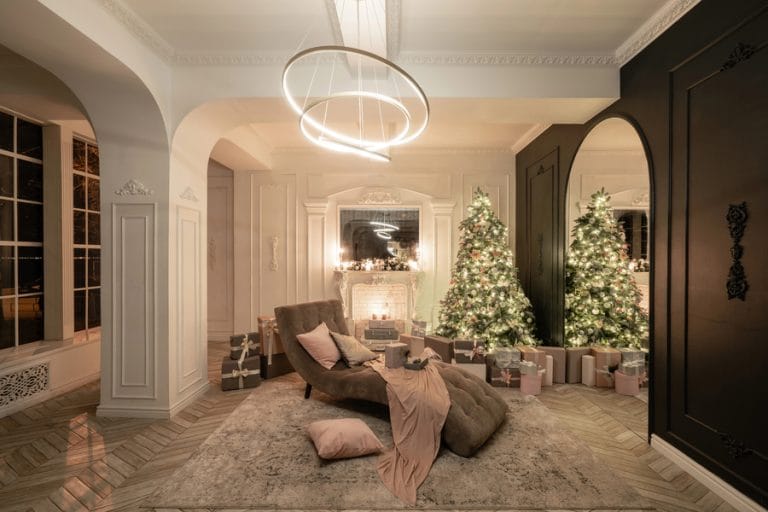 An elegant living room decorated for Christmas with two lit trees, a cozy chaise lounge, wrapped gifts, and modern circular lighting.