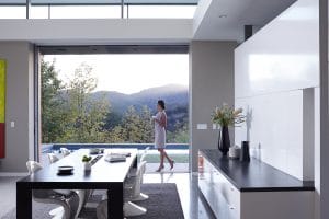 A woman holding a cup walks through a modern open-concept home with large sliding doors revealing a scenic mountain view and a poolside patio.