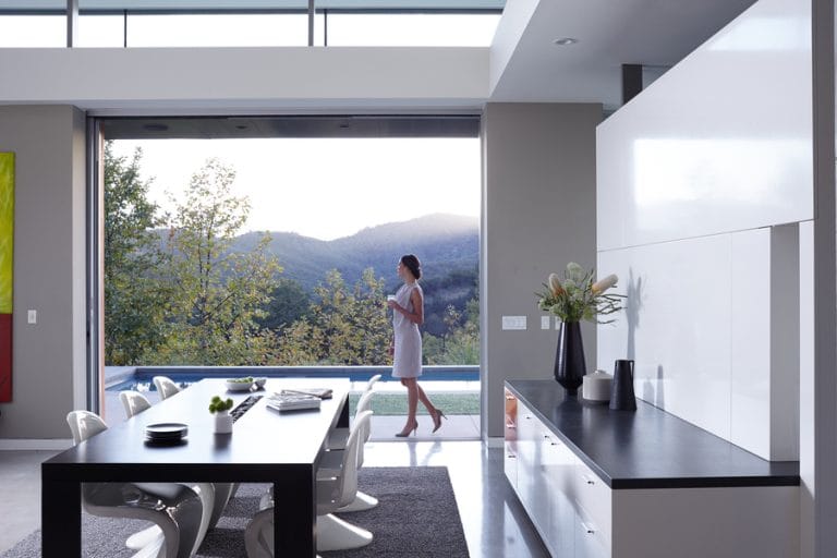 A woman holding a cup walks through a modern open-concept home with large sliding doors revealing a scenic mountain view and a poolside patio.