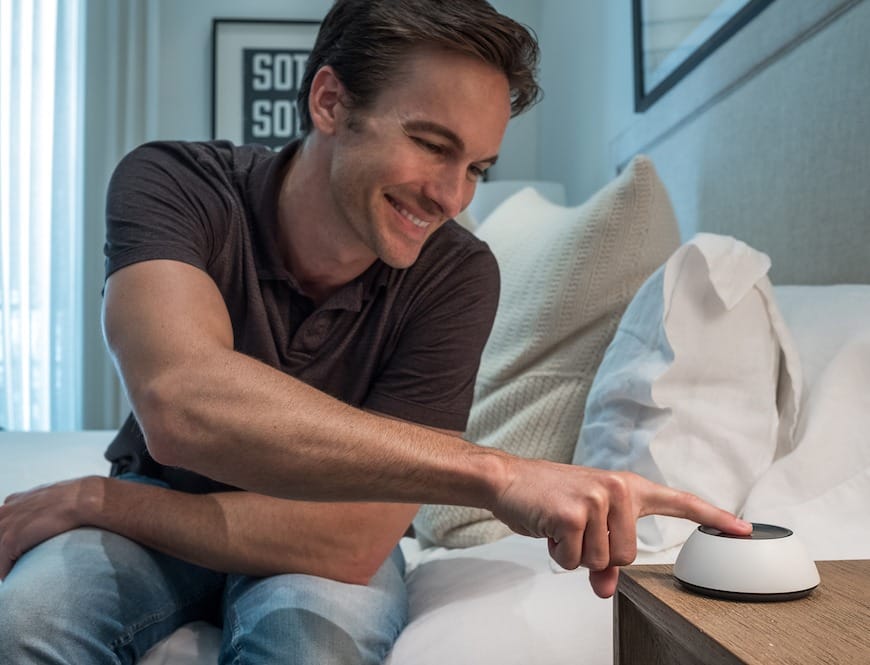 A man sitting in bed, tapping a Josh.ai device on the nightstand.