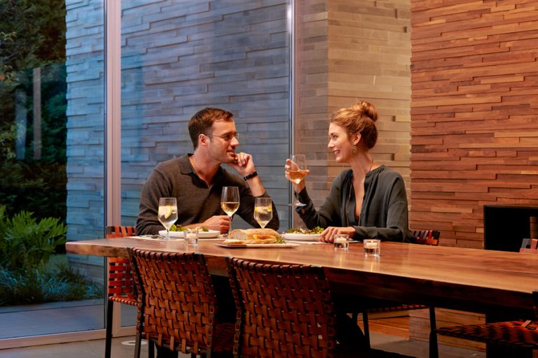 Couple enjoying dinner and wine in a modern dining room with warm lighting, wood accents, and floor-to-ceiling windows.