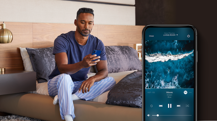 A man looking at the Savant app on his phone. A display to the side shows the Savant app playing music in the living room.